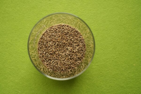 Cumin seeds in a clear glass bowl on a vibrant green background, viewed from above.